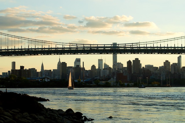 View of sun setting behind a sailboat in the East River behind the Robert F. Kennedy Triborough Bridge and NYC skyline from Astoria Park